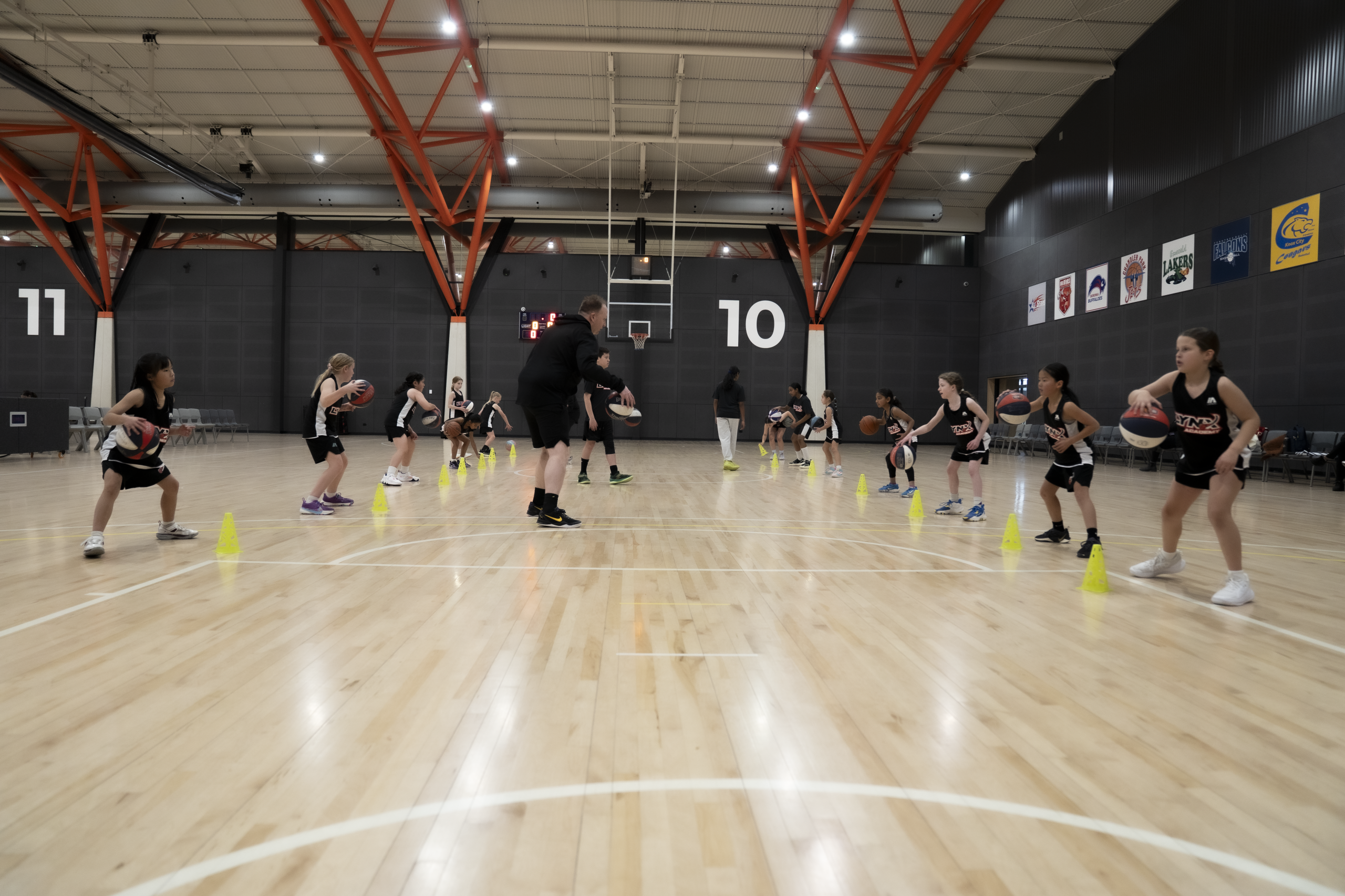 Lynx Academy coaches supervising youth basketball players in black and white jerseys during cone dribbling drills on indoor court with orange beams and numbered walls