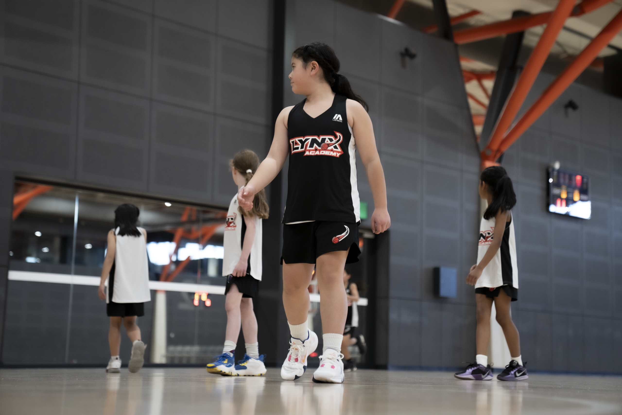 Group of young Lynx Academy basketball players in uniform gathered on indoor court with orange beams and windowed wall during team coaching session